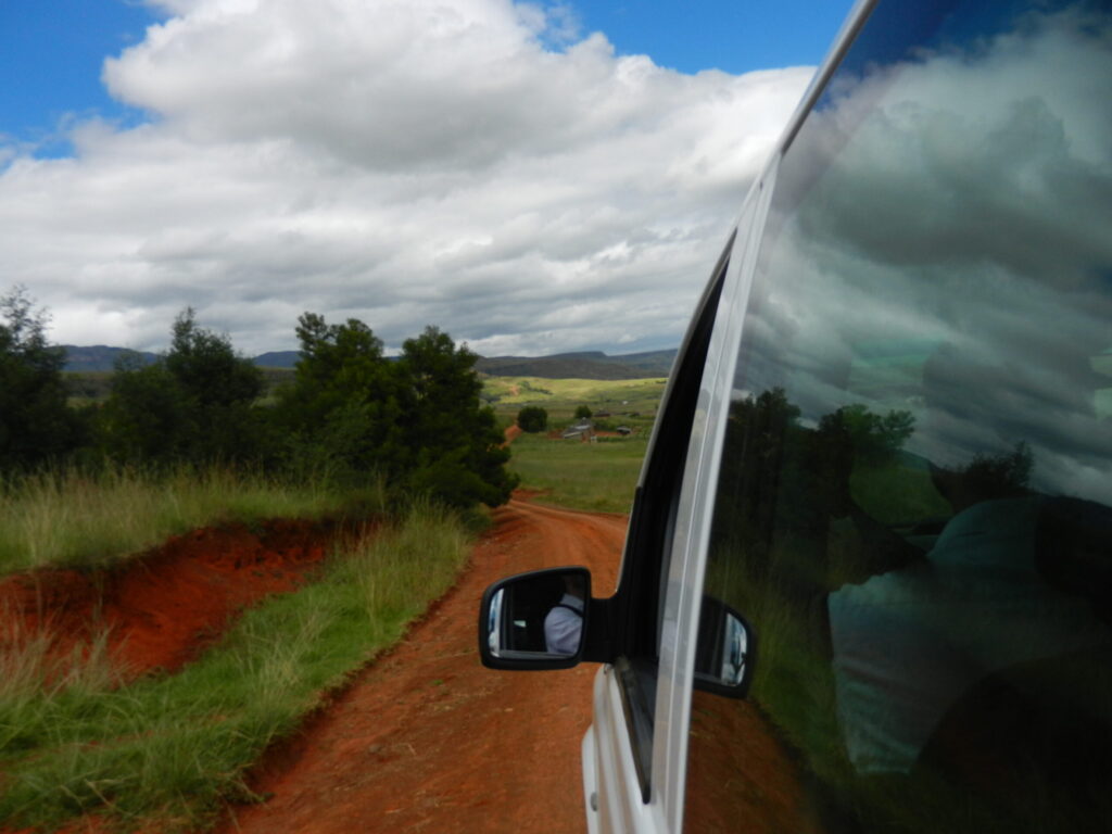 Trekker.space page background image is a Photo by Jason Payne while riding along a dirt road with friends in Kwazulu Natal, South Africa in 2012. The view is out of the window of a trekker mini van and depicts the side of the slowly moving van, blue sky with white cumulous clouds, green vegetation and African grass contrasted with the red/orange clay dirt road that we were traveling along. The is a bend ahead on the road. The trek continues.